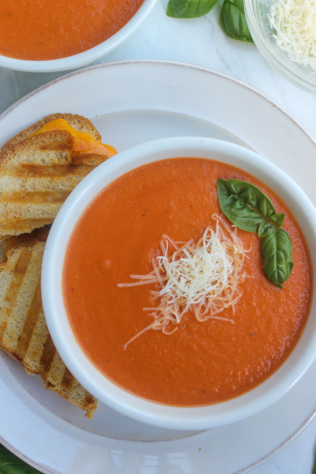 A bowl of White Bean Tomato Soup on a white plate with grilled cheese and a basil leaf.