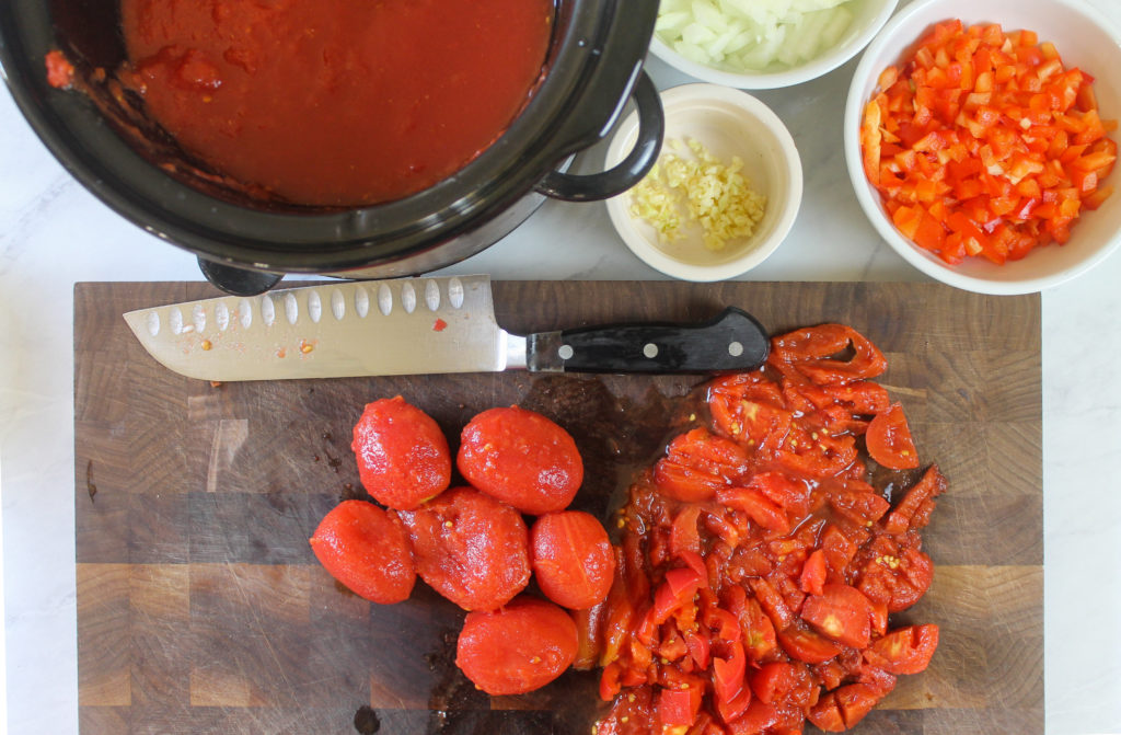 Chopping canned tomatoes on a cutting board and adding to the slow cooker for chili.