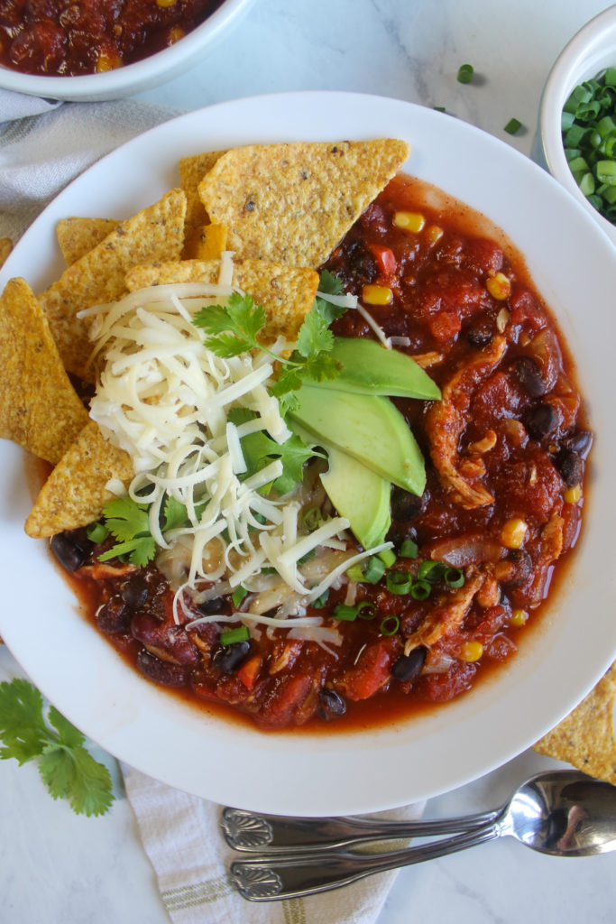 A white bowl of Southwest Chicken Chili topped with shredded cheese, sliced avocado and corn tortilla chips.