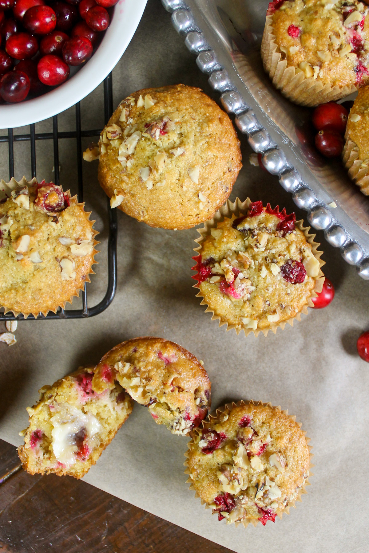 Top down view of cranberry muffins on parchment paper next to a platter and a slice and buttered muffin.