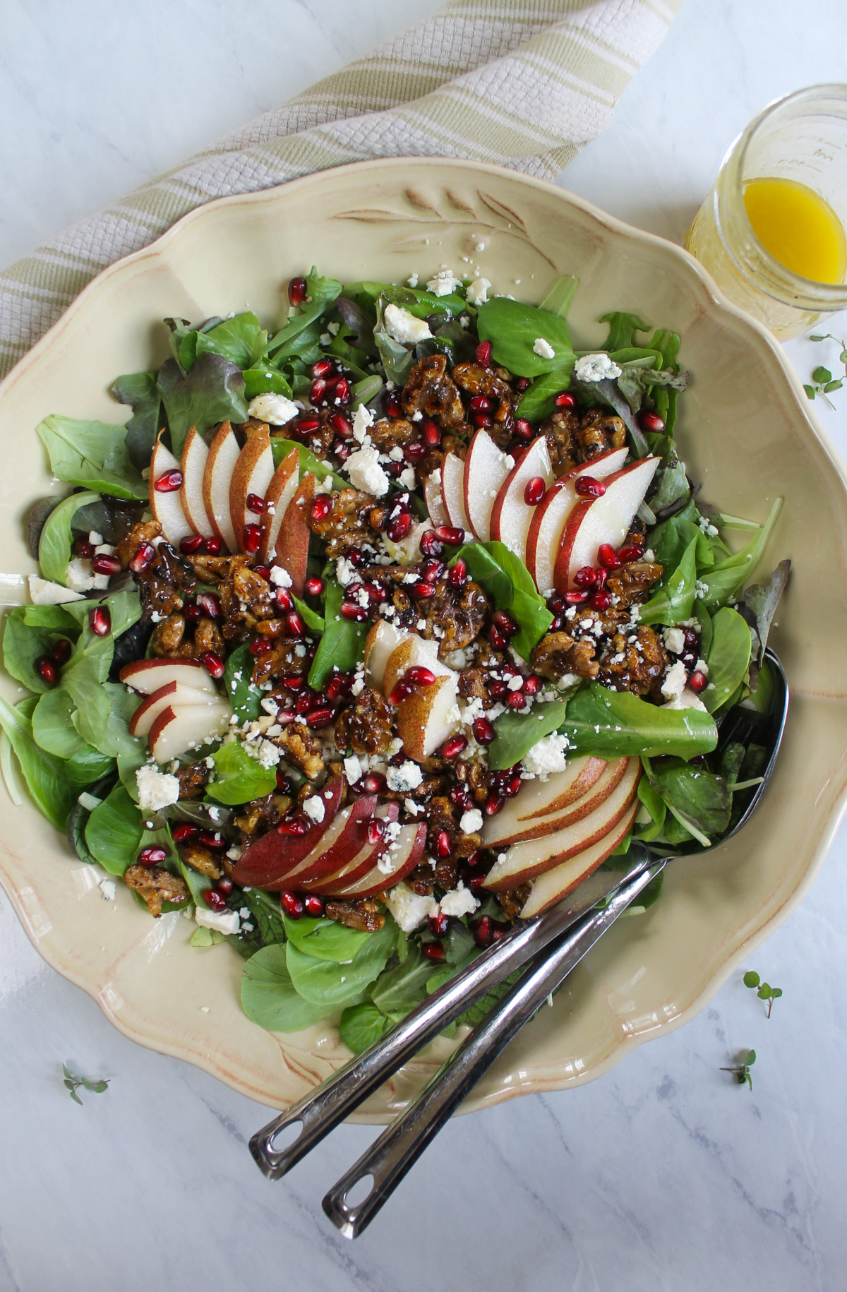 A salad with pear slices, blue cheese crumbles, pomegranate seeds and glazed honey walnuts with a jar of vinaigrette dressing.