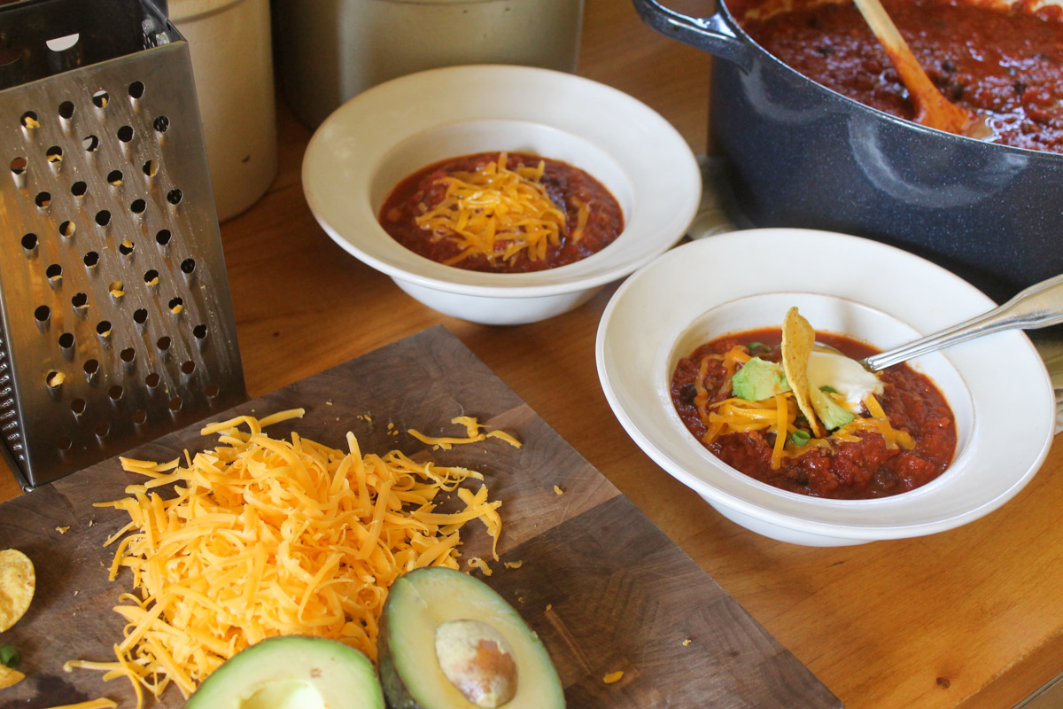Two white bowls of chili next to a cutting board with toppings like shredded cheese and avocado.