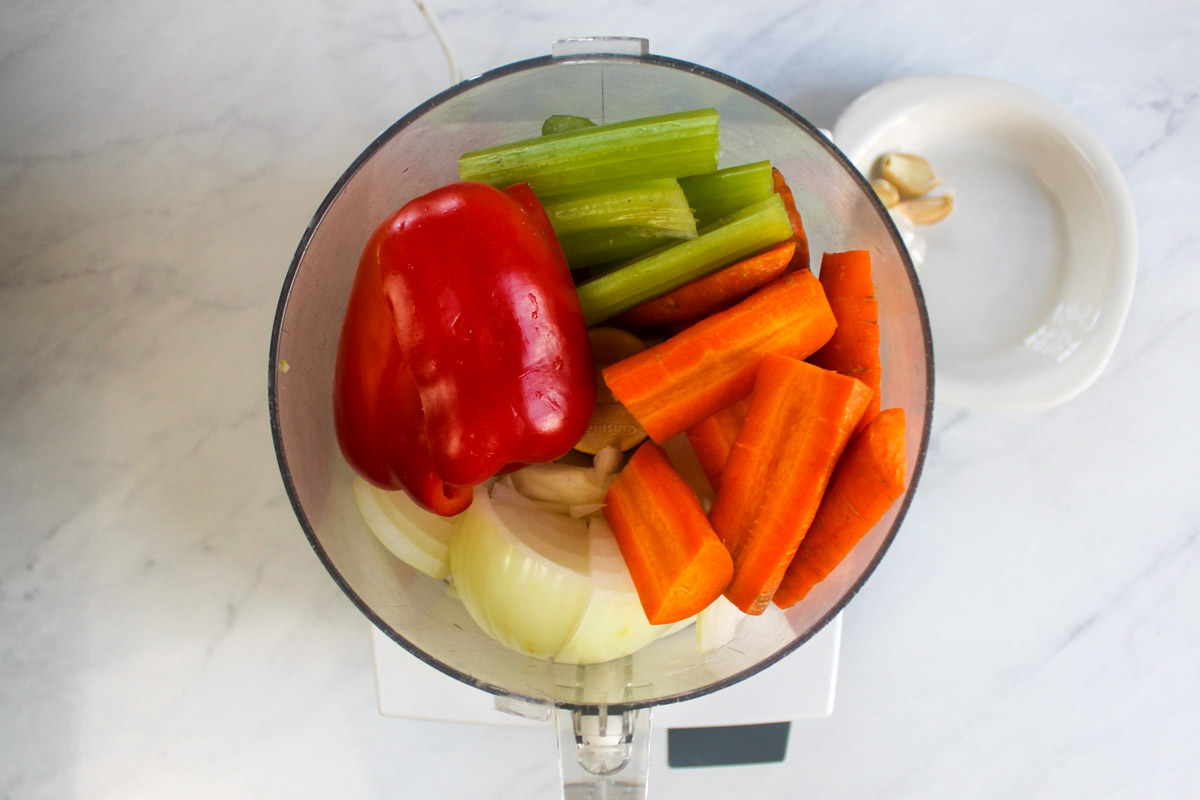 Vegetables in the bowl of a food processor including onion, carrot, celery and red bell pepper.