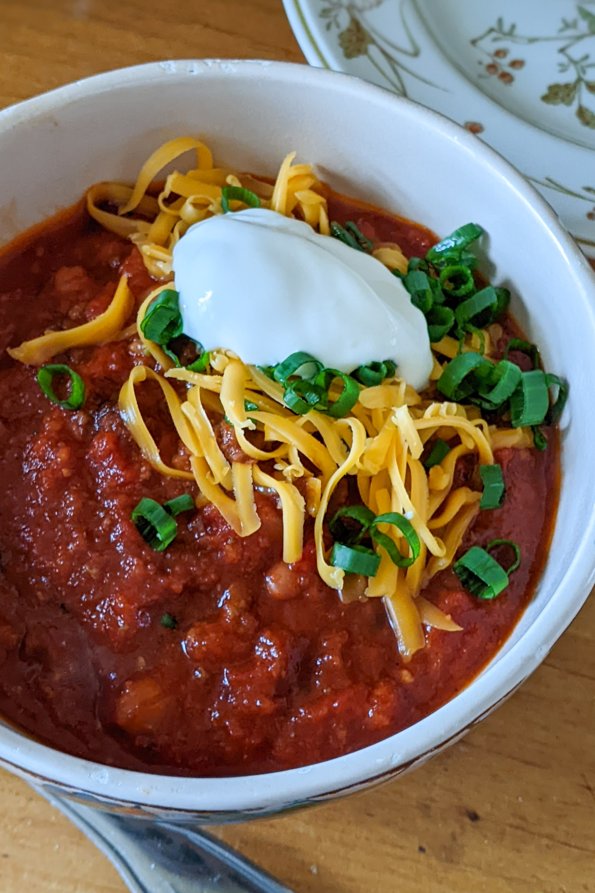 A bowl of ground beef chili topped with shredded cheese, plain yogurt and scallions.