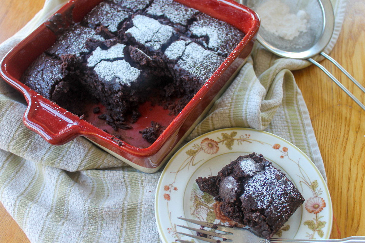 A brownie on a flowered plate in front of a red pan of brownies.