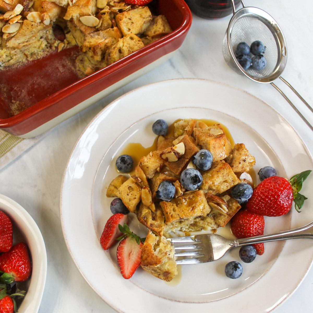 A serving of protein French Toast Bake on a plate next to the casserole with with berries.