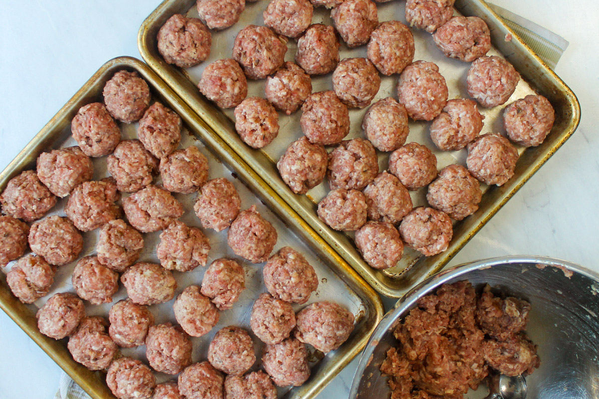 Two large sheet pans of raw meatballs, next to a bowl of meatball mix.