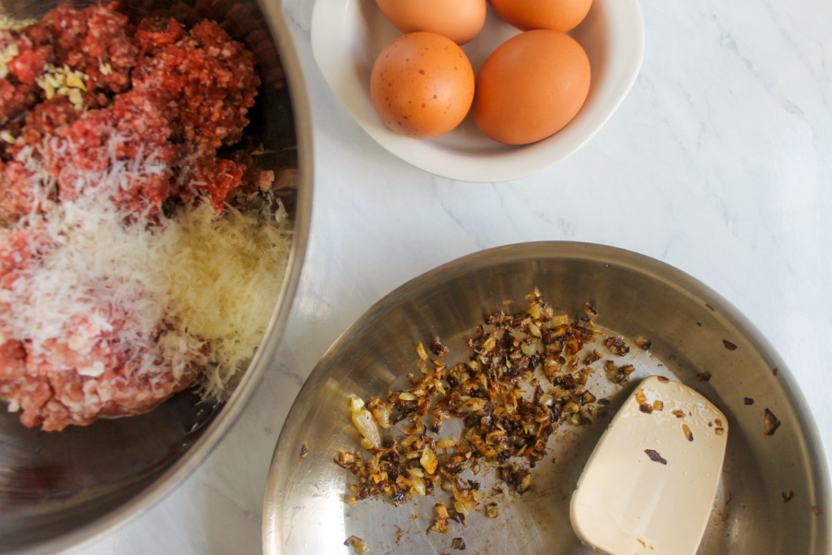 Caramelized onions in a skillet with a bowl of meatball mix and a dish of brown eggs.