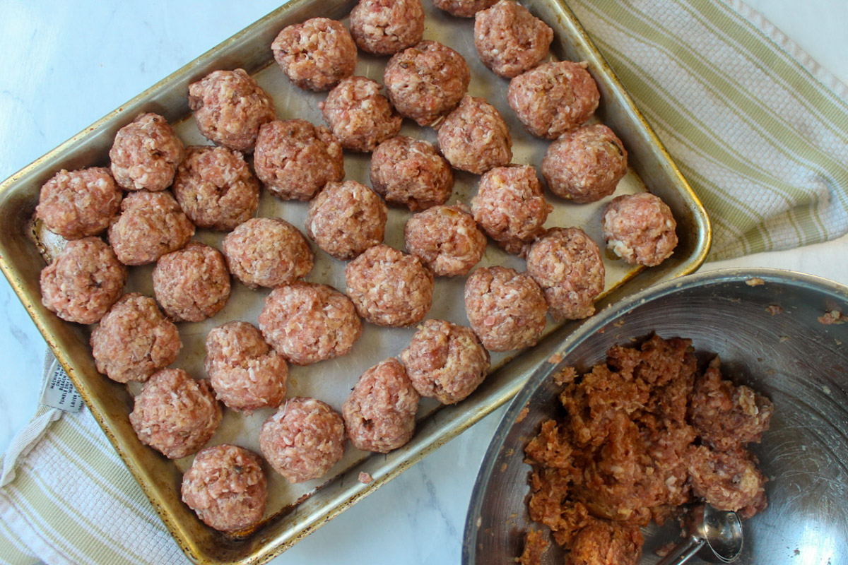 Raw meatballs on a sheet pan next to a bowl with a portion scoop.