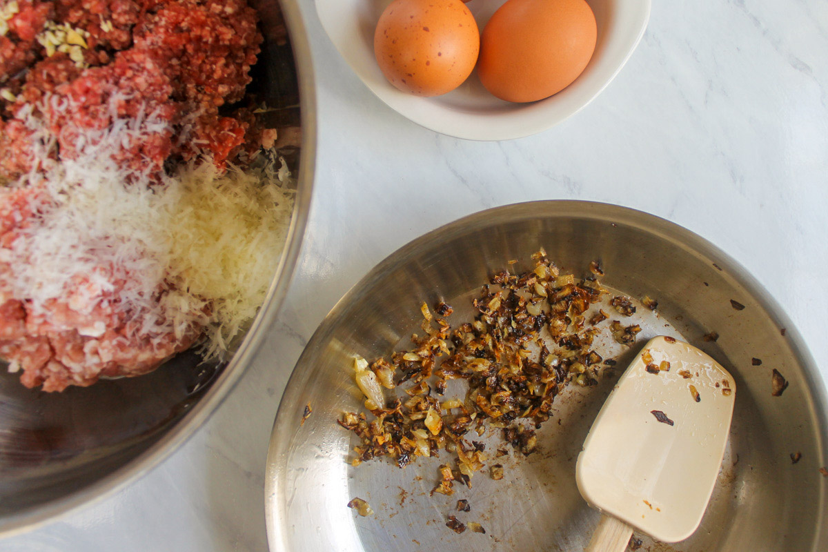 Caramelizing onions in a skillet next to a bowl of meatball mixture and two brown eggs.