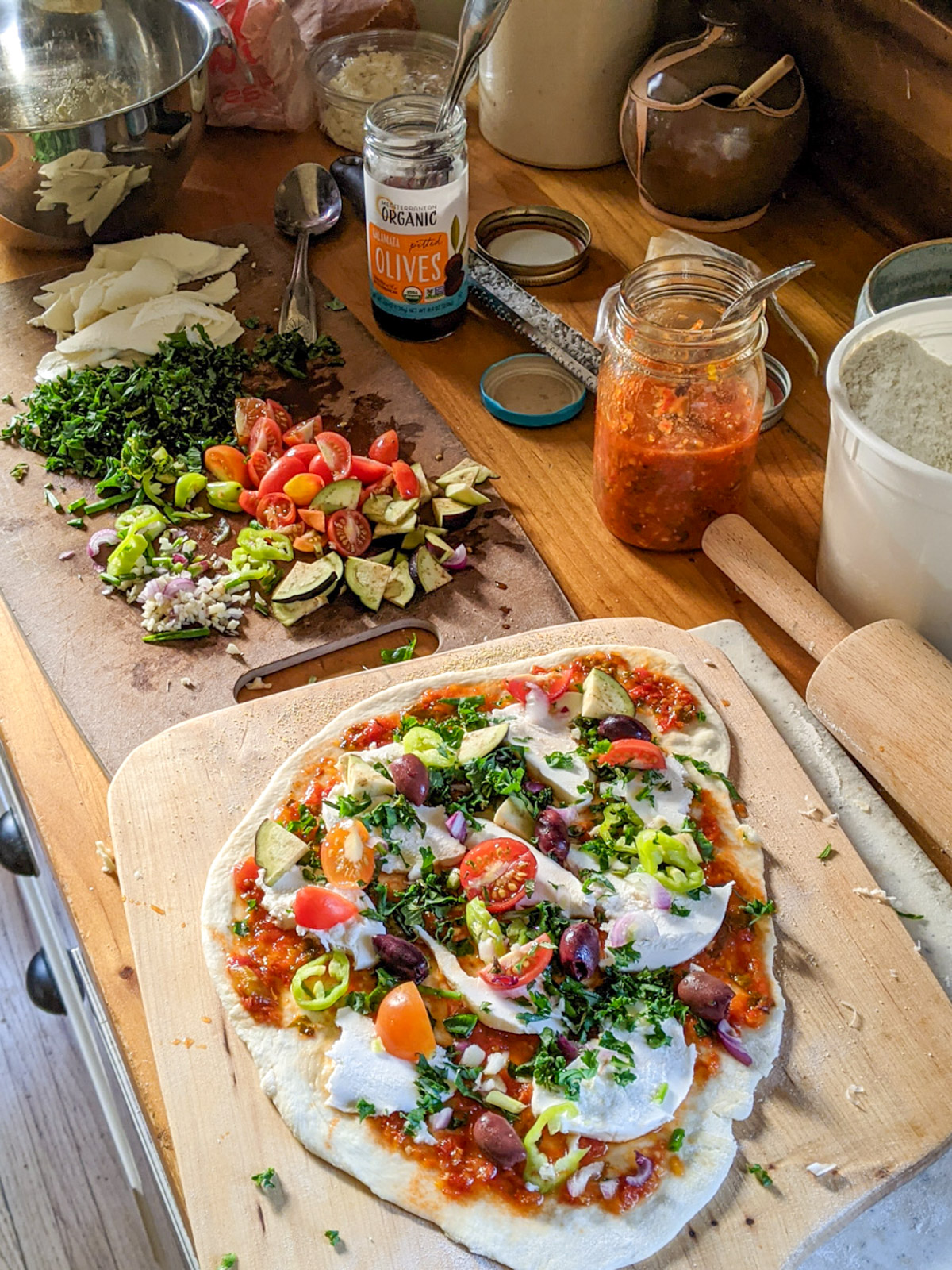 Veggie pizza toppings prepped on a cutting board with a pizza ready to go into the oven.