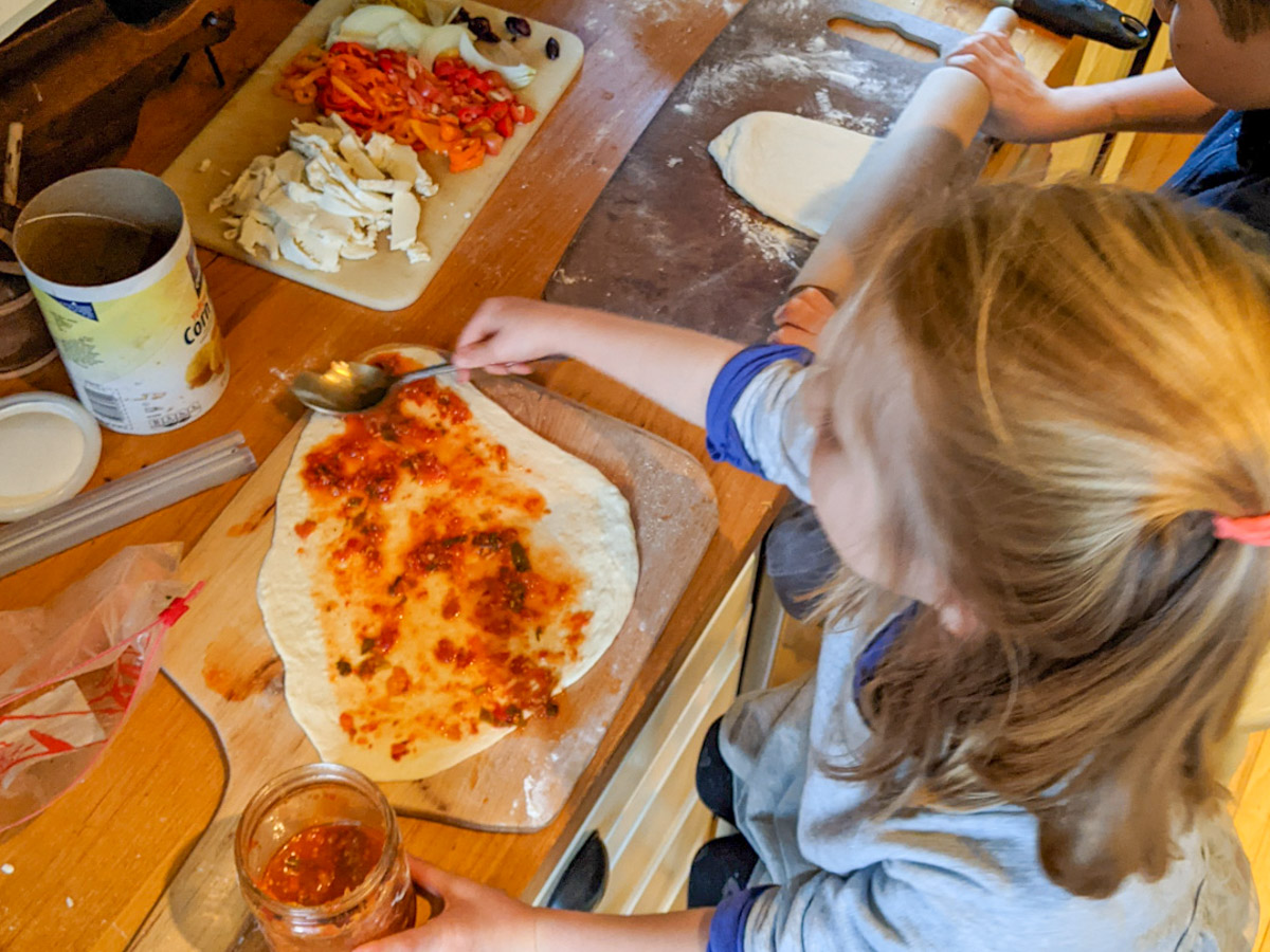 Kids making homemade pizza, adding pizza sauce to rolled out dough.