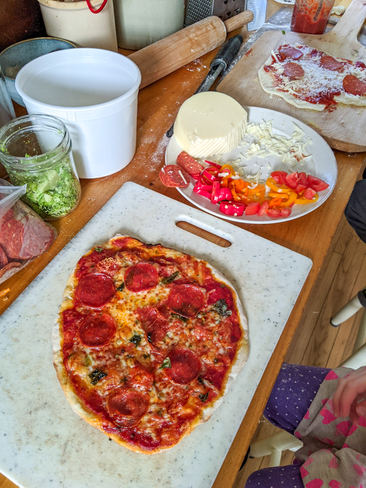 Pizza making on the countertop, a finished pizza and a plate of shredded cheese and toppings.