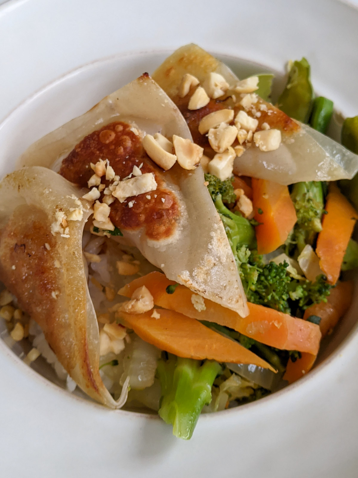 A white bowl of stir fry rice and vegetables topped with 3 chicken apple potstickers and crushed peanuts.