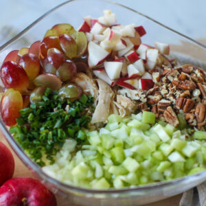 A glass bowl of ingredients for Chicken Salad with grapes, apples, pecans and celery.