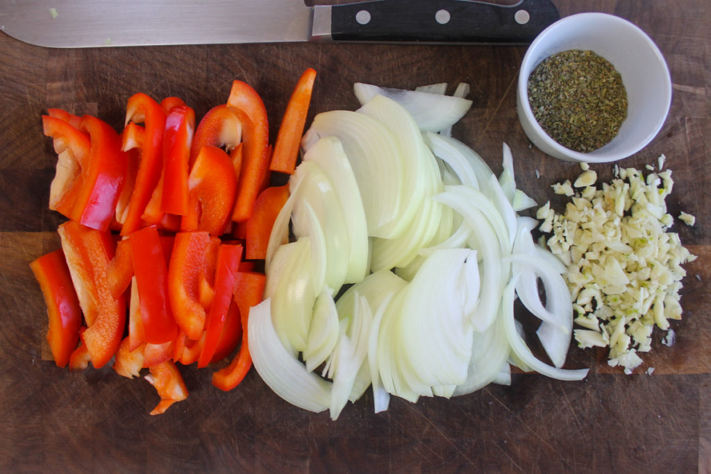 Sliced red bell pepper, onion and minced garlic on a cutting board with knife.