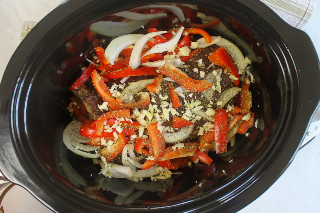 Italian beef roast being prepared in the slow cooker with onions, peppers and garlic.