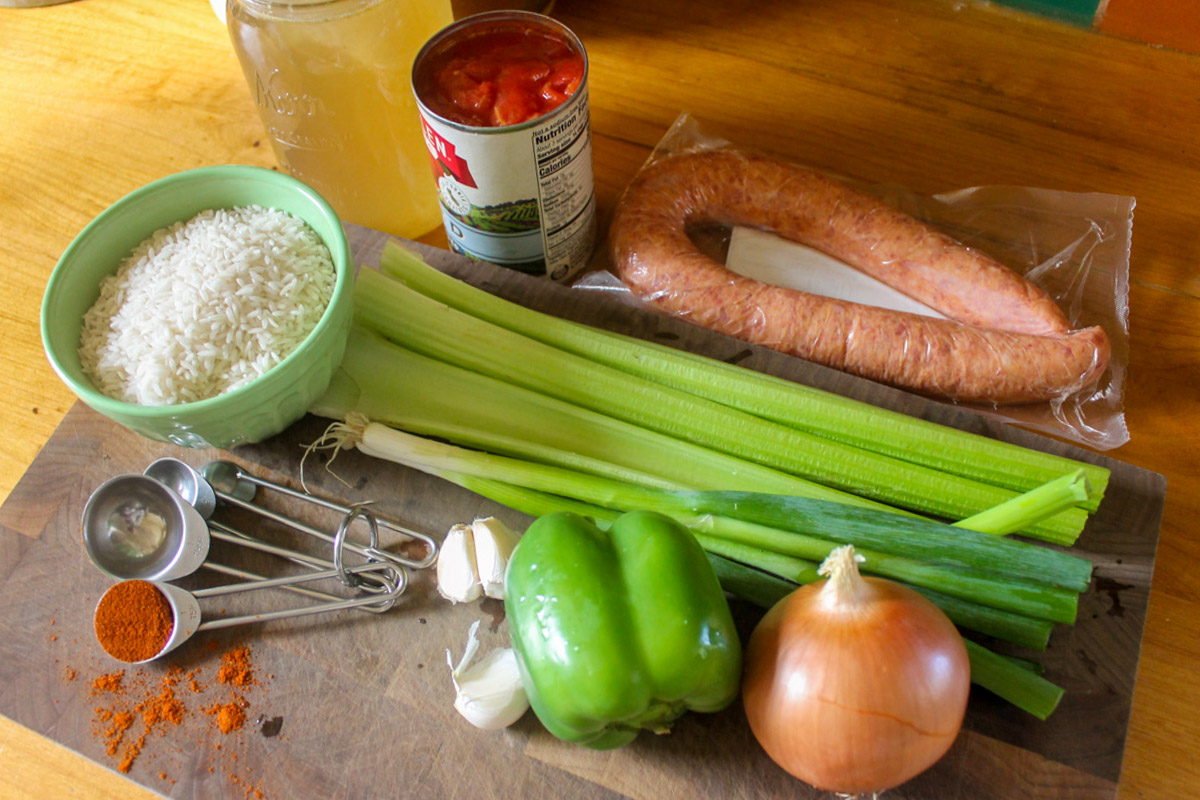 Ingredients for jambalaya on a cutting board including celery, green pepper, onion, rice, sausage, canned tomato and chicken stock.