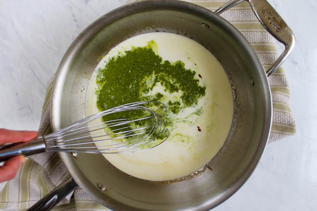 Homemade pesto and heavy cream in a skillet with a whisk to make Pesto Alfredo Sauce.