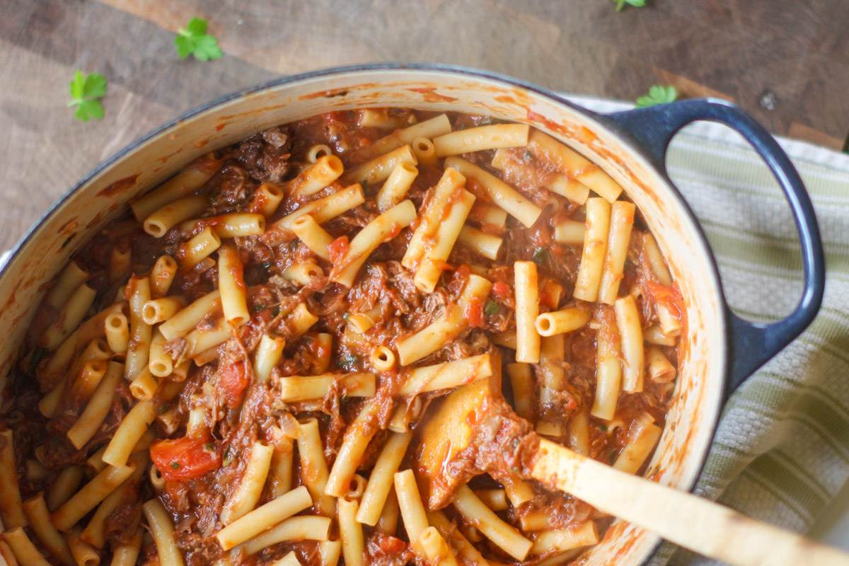 A pot of ragu sauce with leftover shredded beef and ziti noodles mixed in with a wooden spoon.