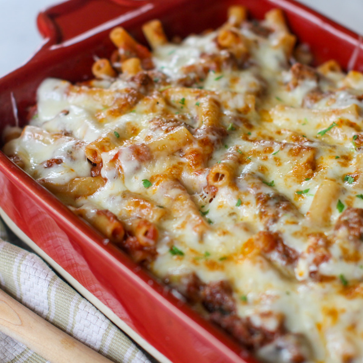 A red baking dish of ziti pasta in a marinara beef ragu sauce baked with melted white cheese.