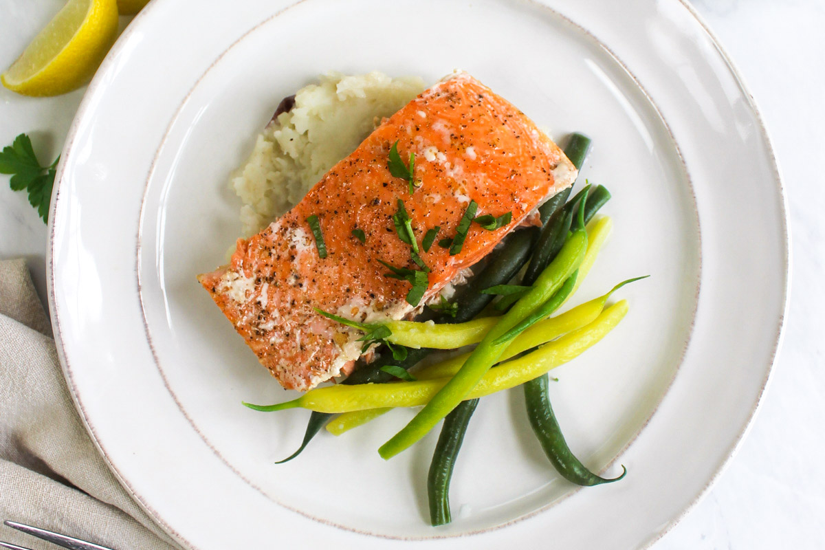 A white plate with a piece of roasted salmon over mashed potatoes with green beans and topped with herbs.