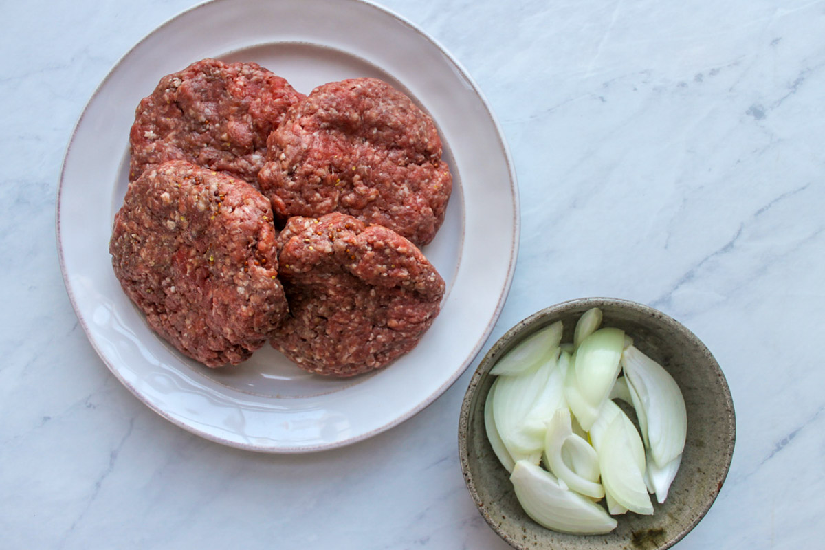 Prepped raw burger patties on a plate and a bowl of sliced raw onions.