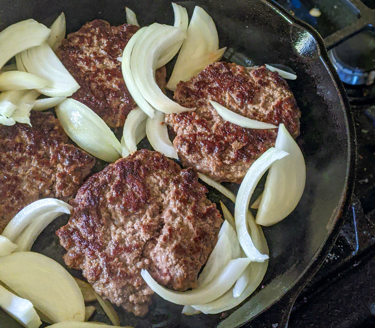 Burgers frying in a cast iron skillet with raw onions added around them to cook.