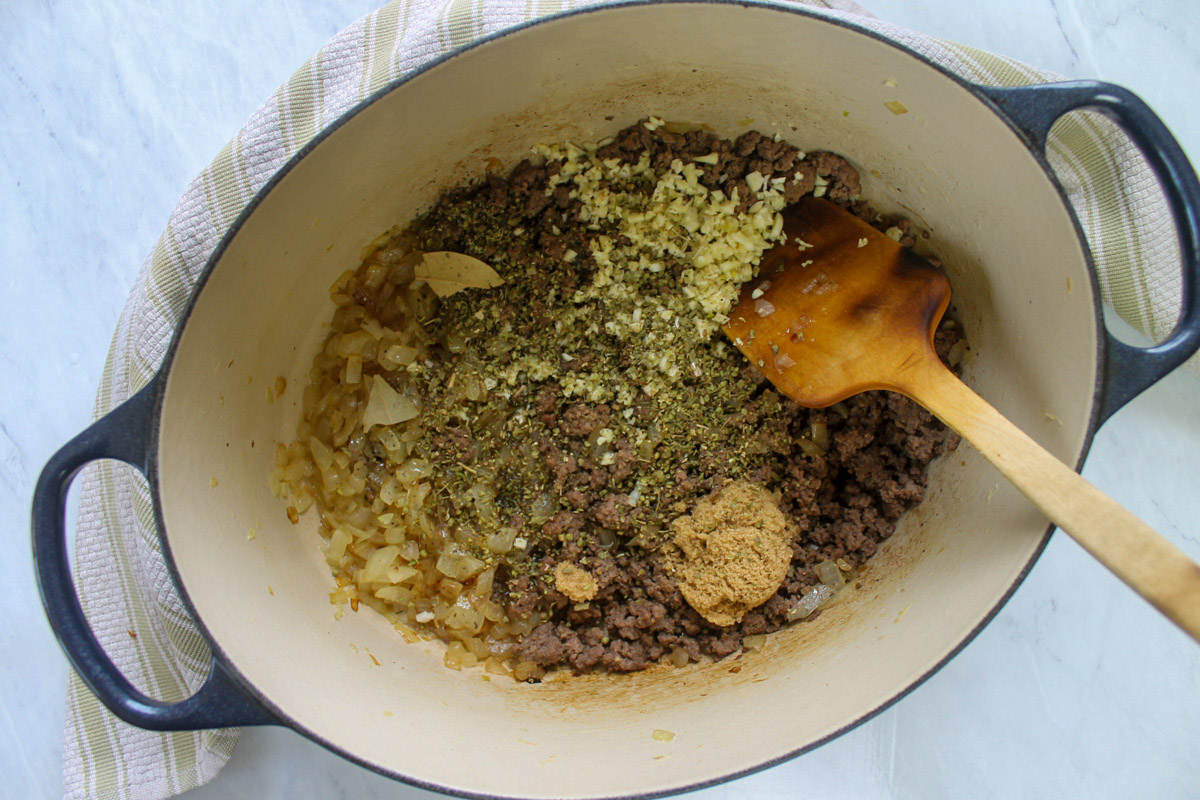 Adding seasonings to the browned ground beef and onions in a pot with a wooden spoon.