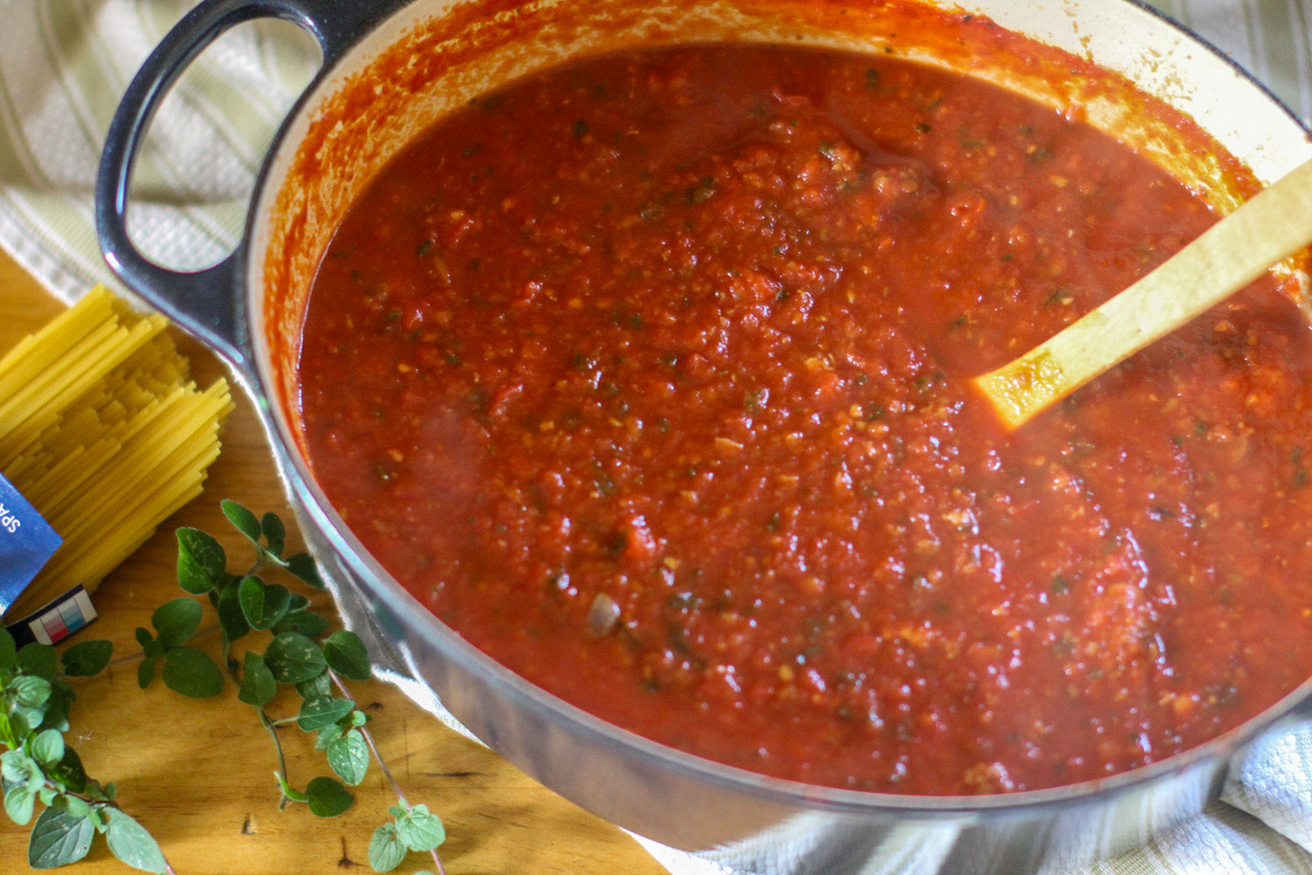 A finished pot of red marinara spaghetti sauce with a box of pasta and herbs next to it.