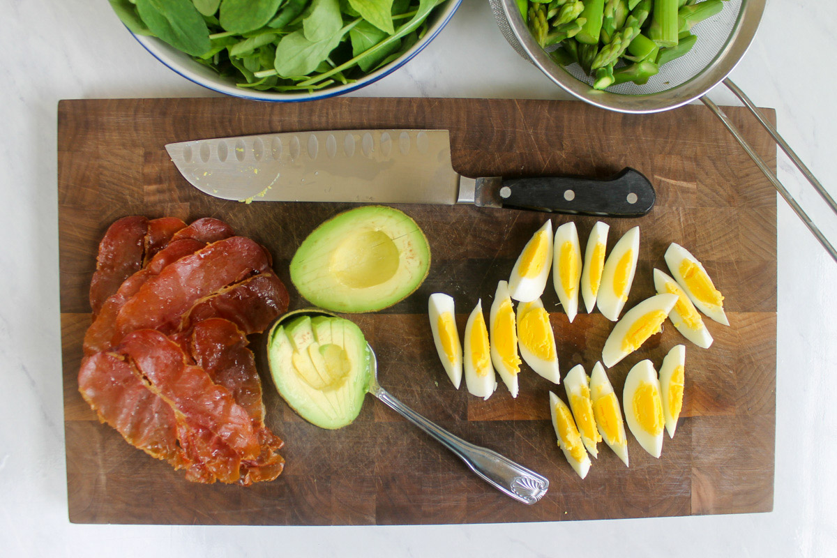 A cutting board with prepared ingredients including hard boiled eggs, sliced avocado and crispy prosciutto.
