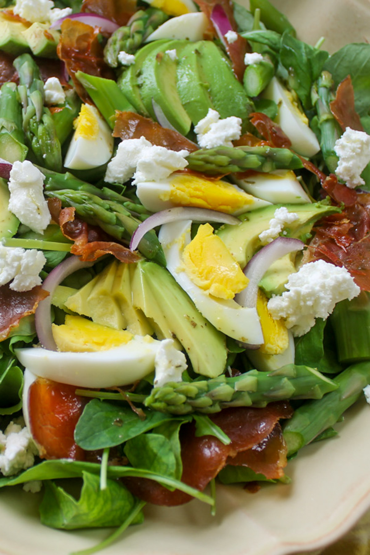 A close up of asparagus salad with sliced avocado, crumbled crispy prosciutto and hard boiled egg slices.