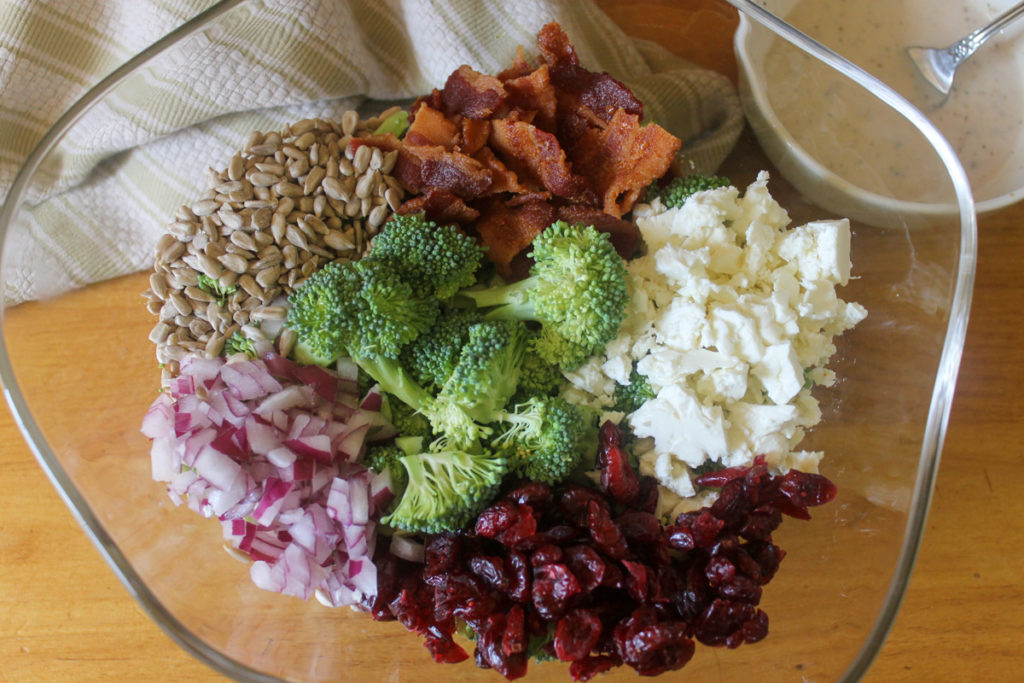 Broccoli Salad ingredients in a bowl, bacon, sunflower seeds, feta, onion, craisins.