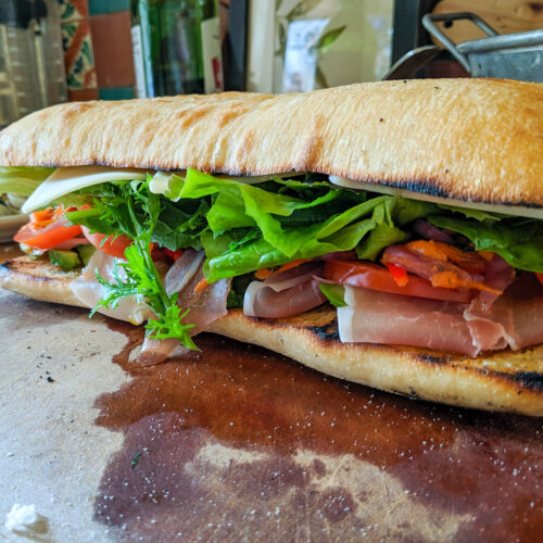 A large sub on ciabatta bread with prosciutto, lettuce, and homemade giardiniera on a cutting board.