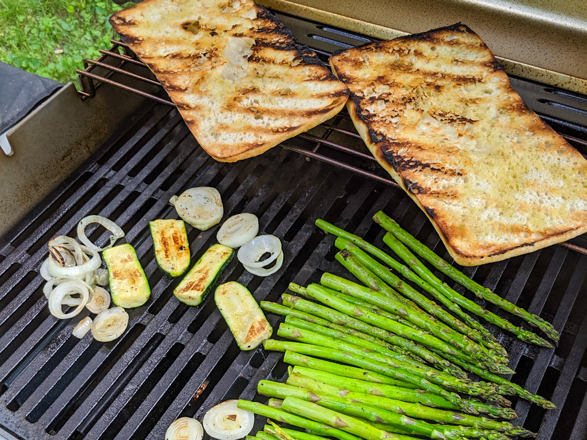 A loaf of Ciabatta bread slice open on a grill with veggies as a side dish.
