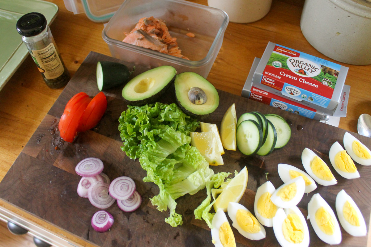 A cutting board with prepared ingredients including sliced hard boiled eggs, avocado, sliced red onion, lettuce and leftover roasted salmon.