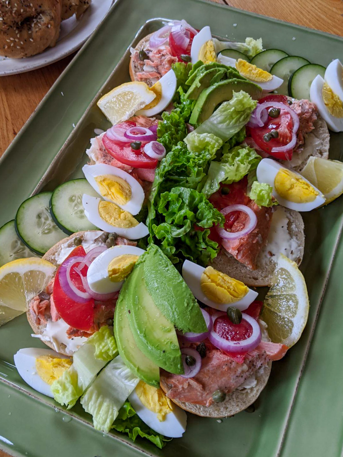 A green platter of bagels topped with cream cheese, cooked salmon, avocado, lemon, egg, lettuce, red onion and capers.