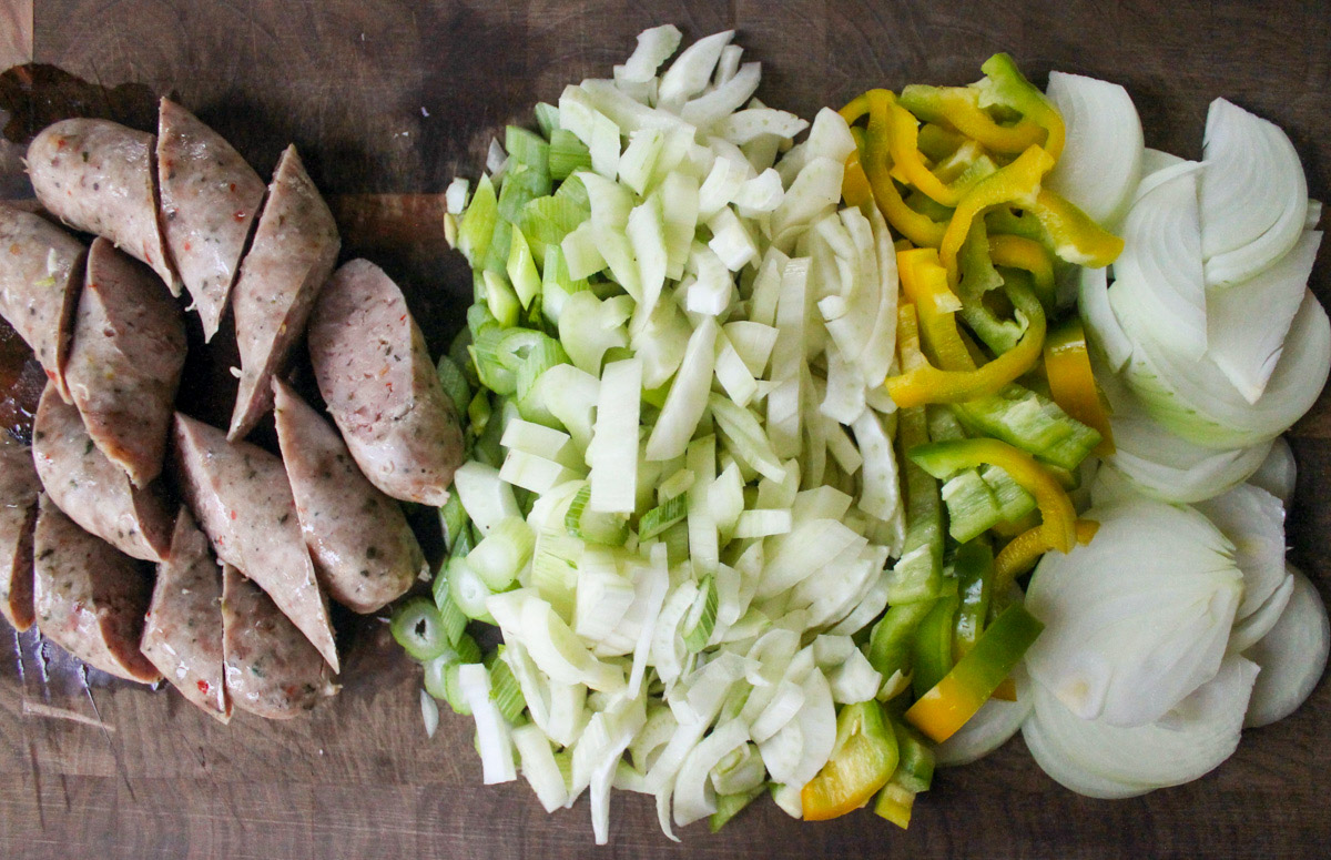 Ingredients, sliced onion, bell peppers, fennel and sausage links on a cutting board.