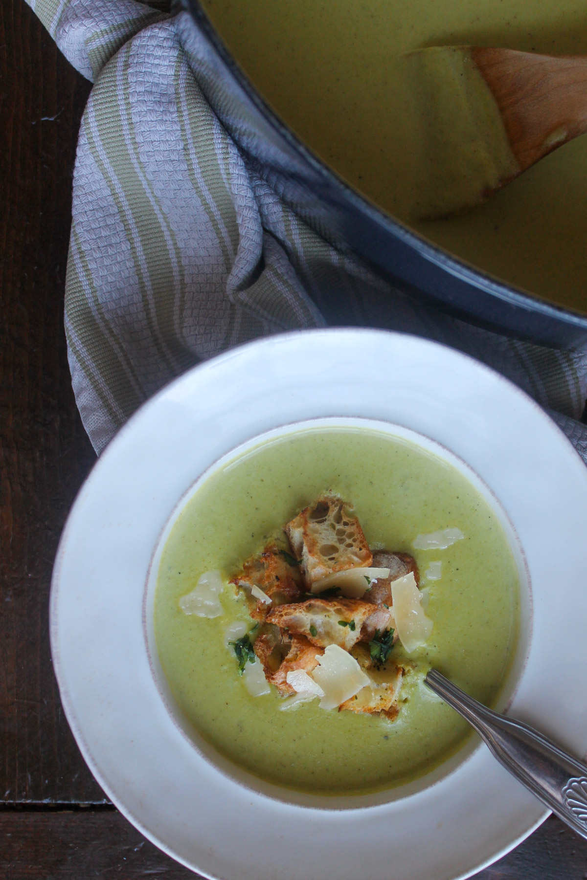 A white bowl of asparagus soup next to a blue Dutch oven pot of soup.