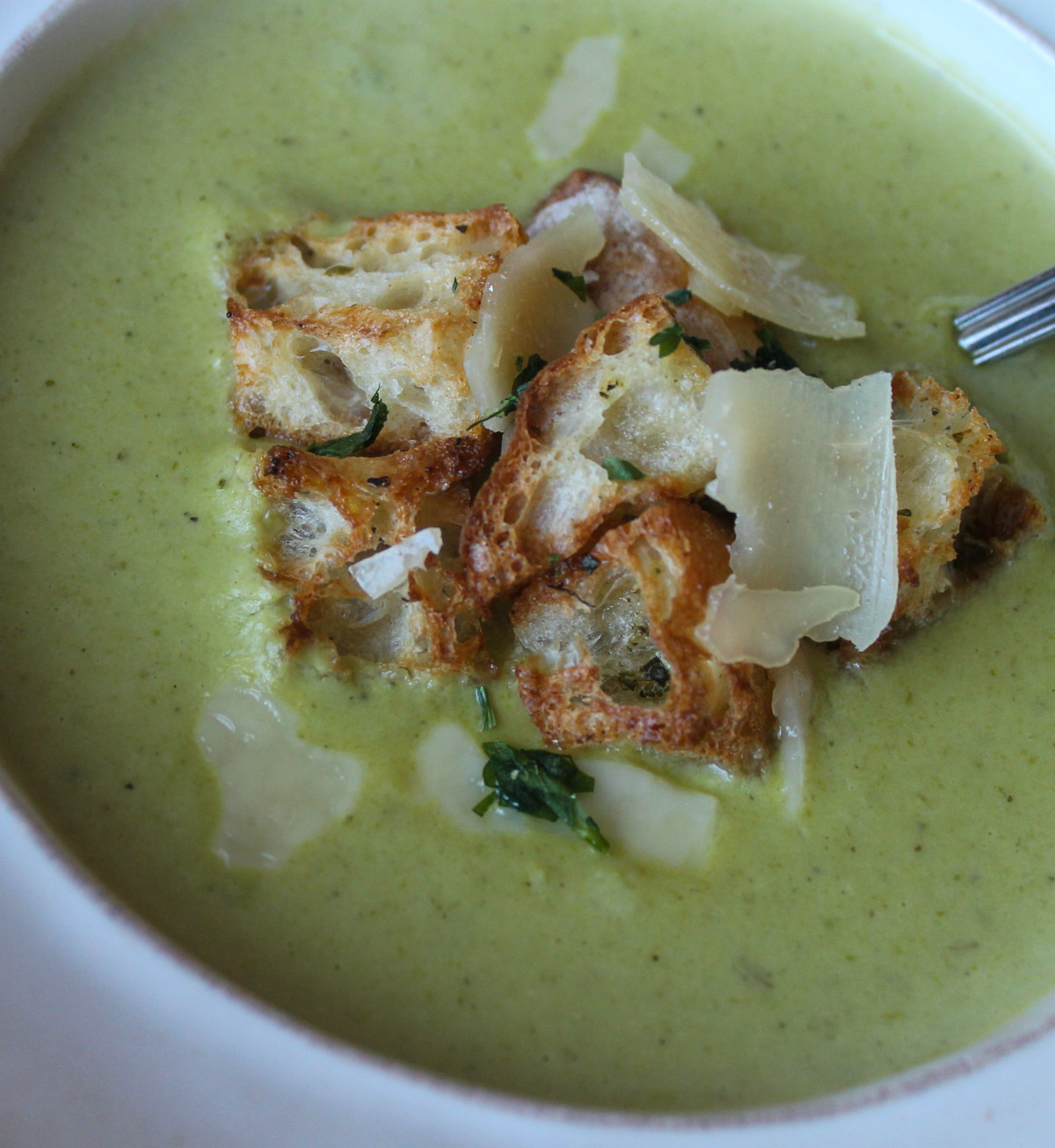 A close up of a bowl of green blended asparagus soup with croutons and Parmesan cheese.