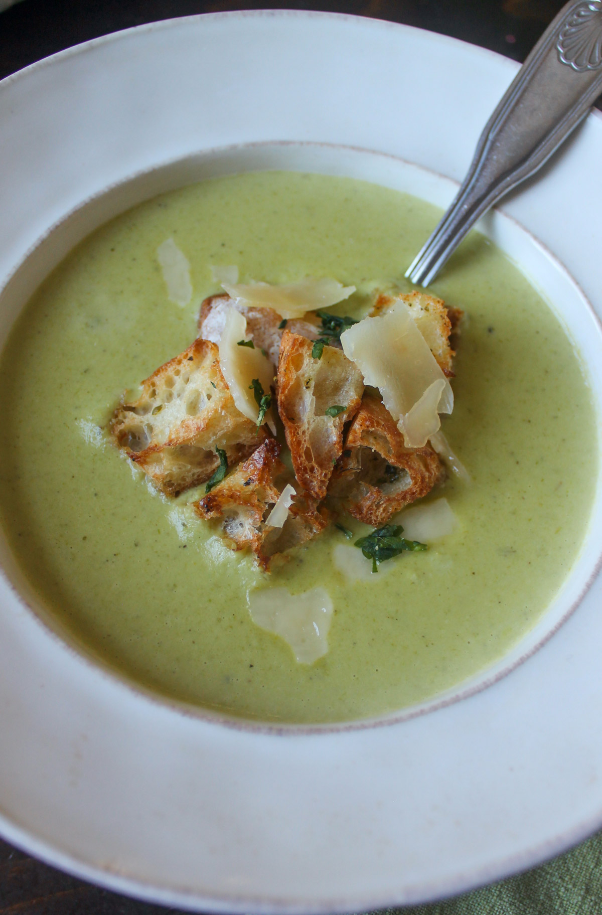 A white bowl of creamy asparagus soup with homemade croutons and Parmesan.