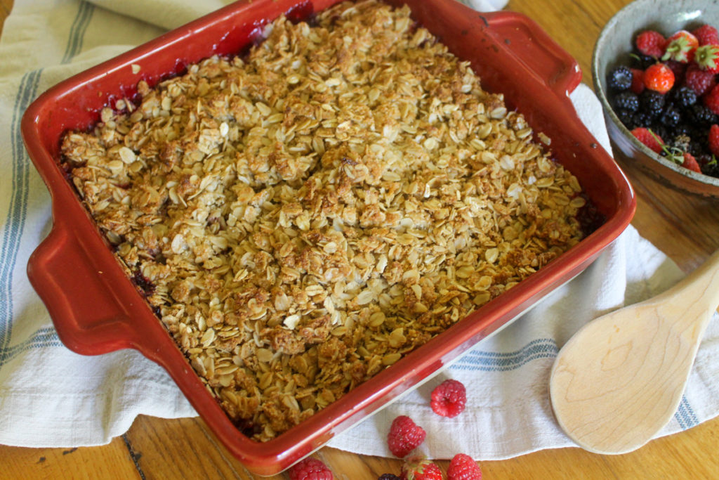 A red baking dish of finished berry rhubarb crisp with a wooden spoon and a bowl of berries.
