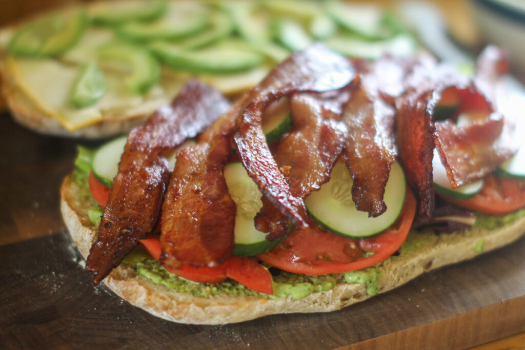 Assembling a Pesto BLT on ciabatta bread with bacon, lettuce, tomato. cucumber and cheese.