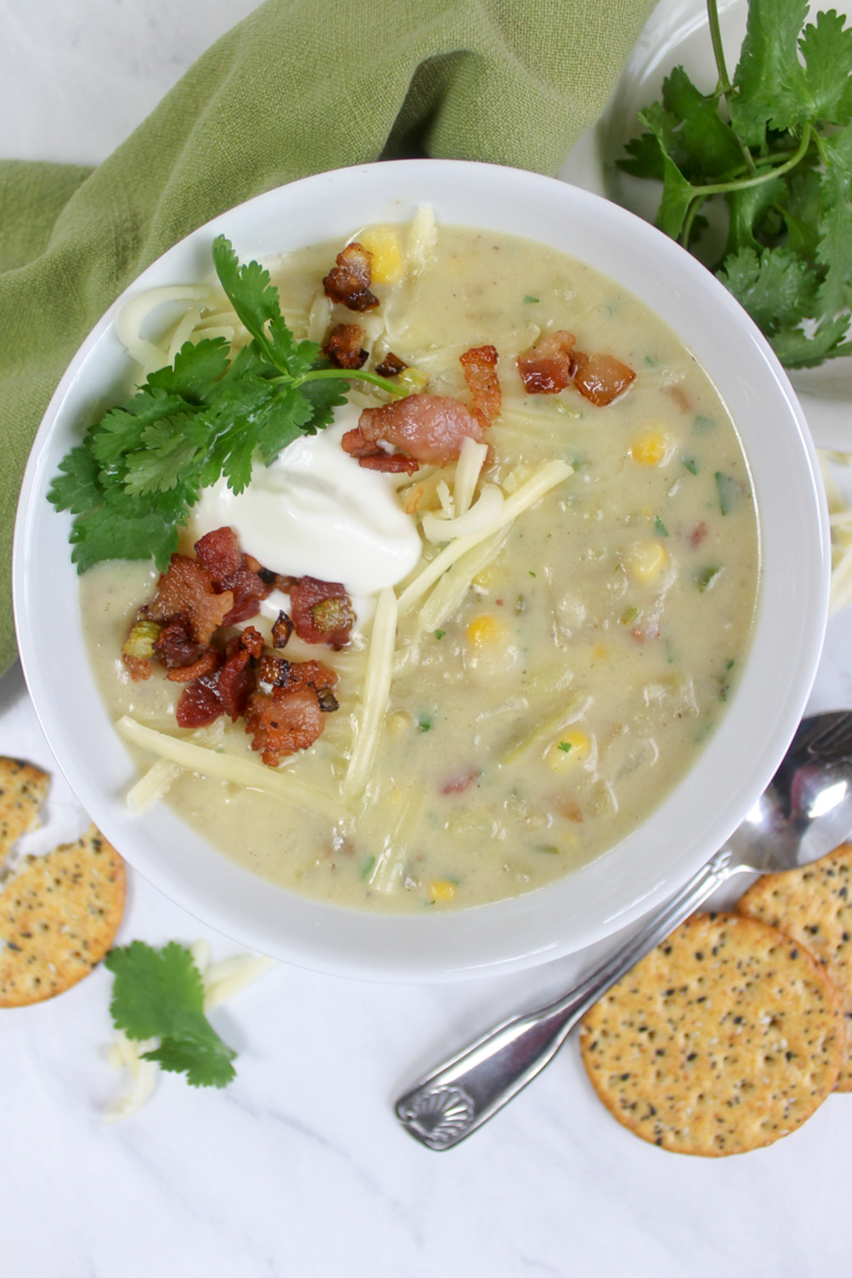 A white bowl of potato corn chowder topped with bacon, plain yogurt, shredded white cheddar and cilantro.