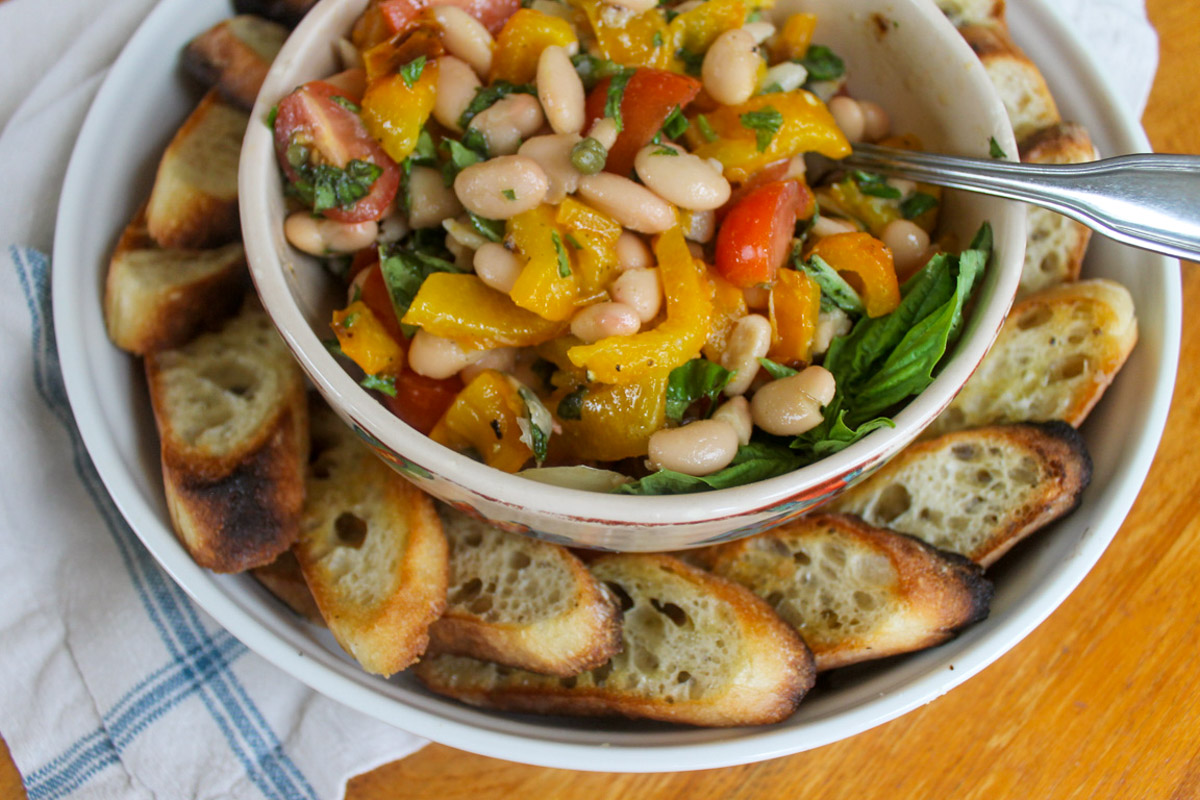 A bowl of white bean bruschetta with roasted pepper and tomato, with crostini around the plate.