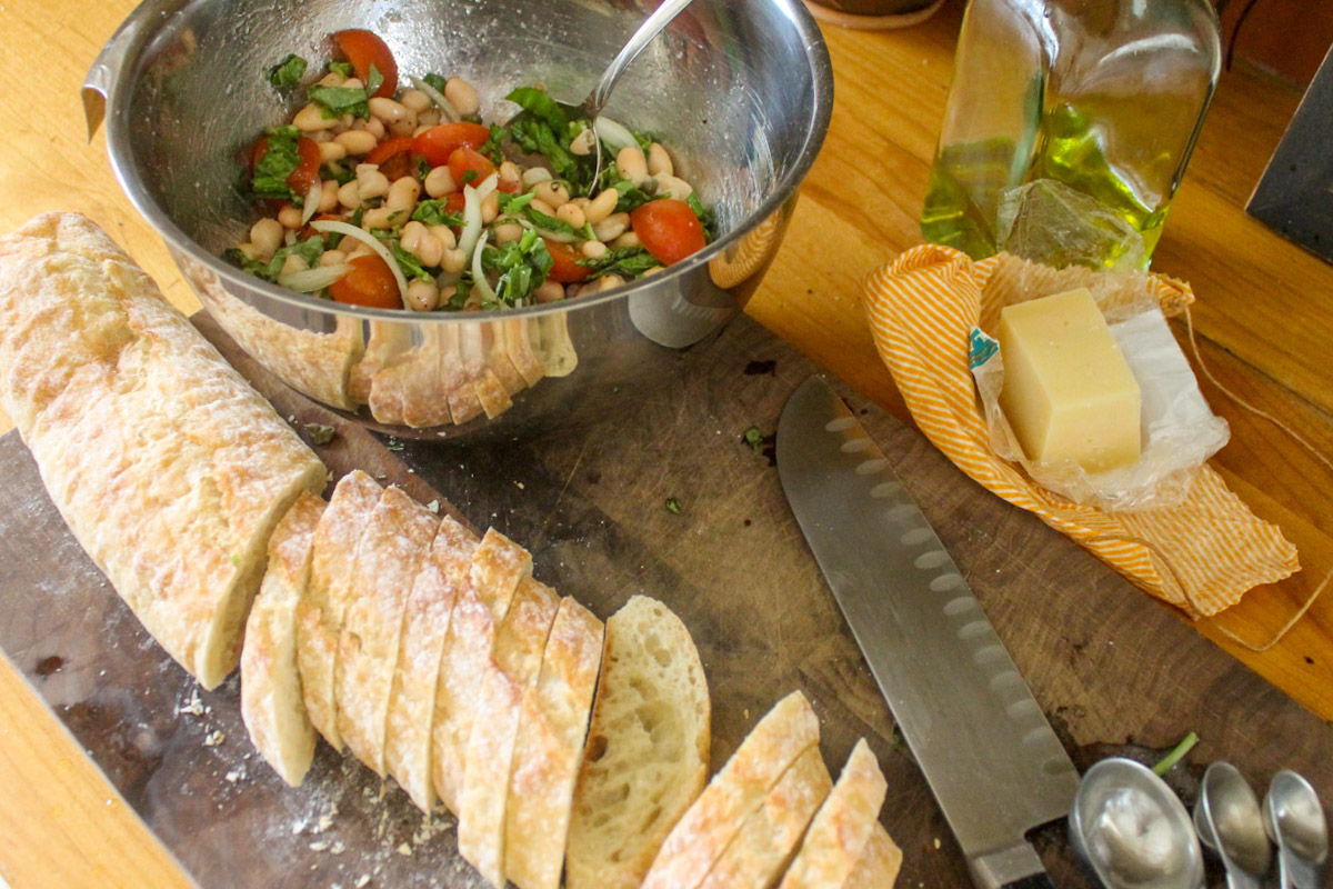 Preparing white bean bruschetta in a bowl, and slicing baguette to make crostini.
