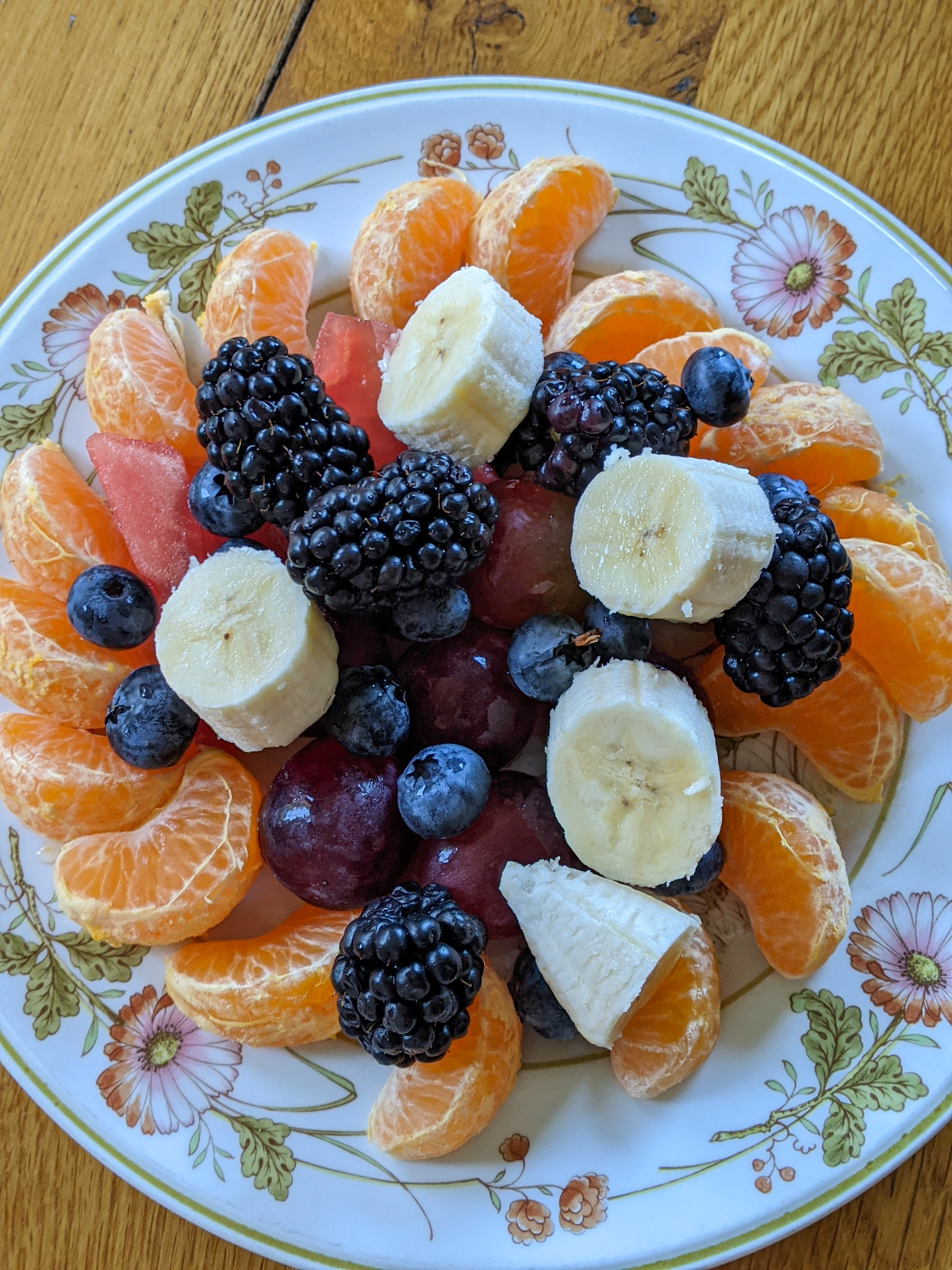 A plate of cutie oranges, blackberries, banana, watermelon, grapes and blueberries.