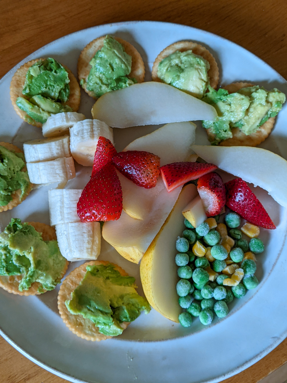 Healthy snacks for kids including mashed avocado on crackers, banana, strawberry, pears, and frozen peas and corn.