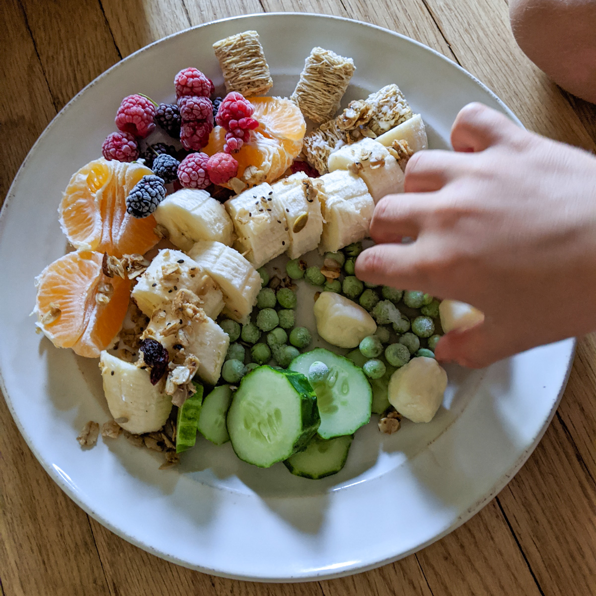 A kid's hand grabbing healthy snacks from a shared plate of fruit, vegetables, and cereal.
