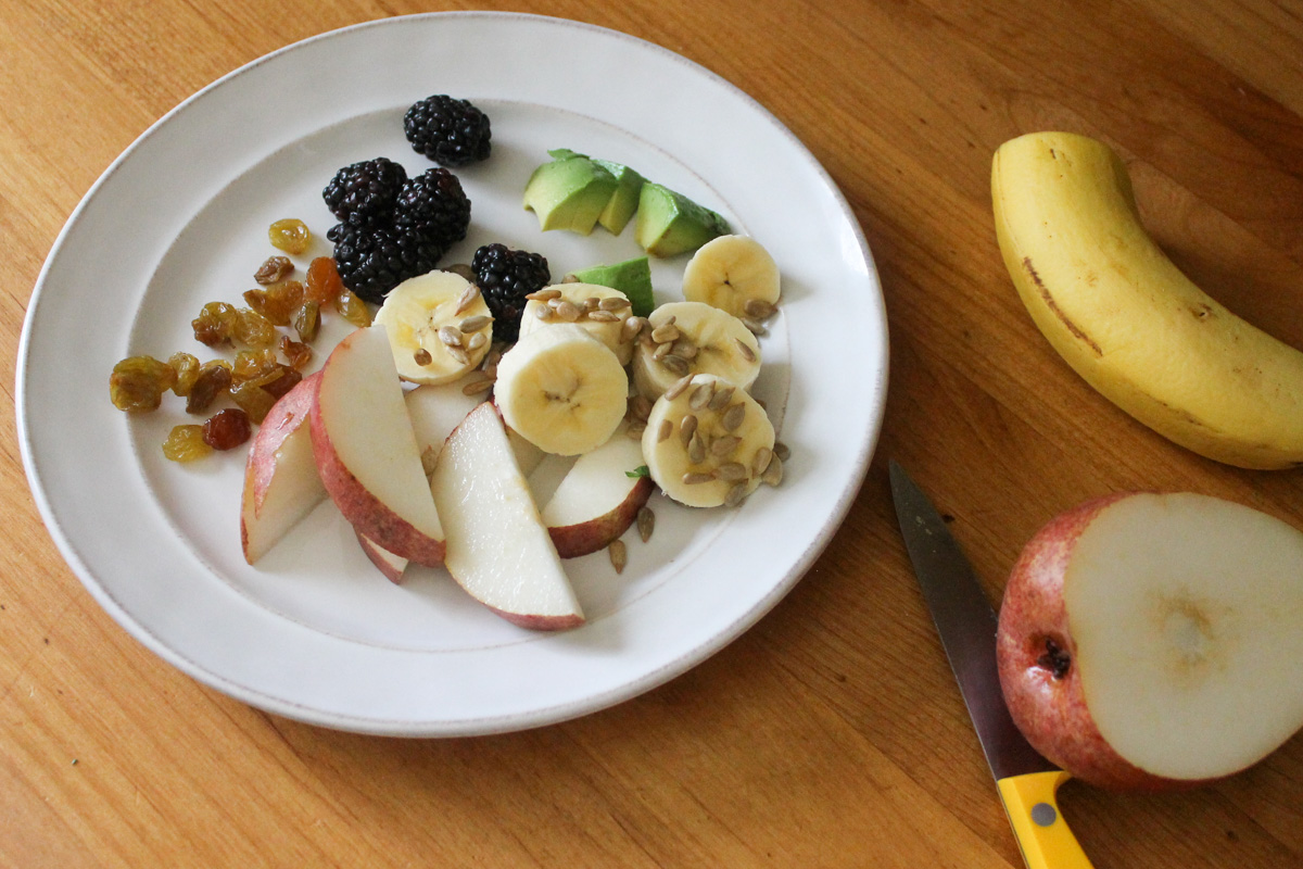 A plate of banana, apple, avocado, black berries and raisins for the kids.